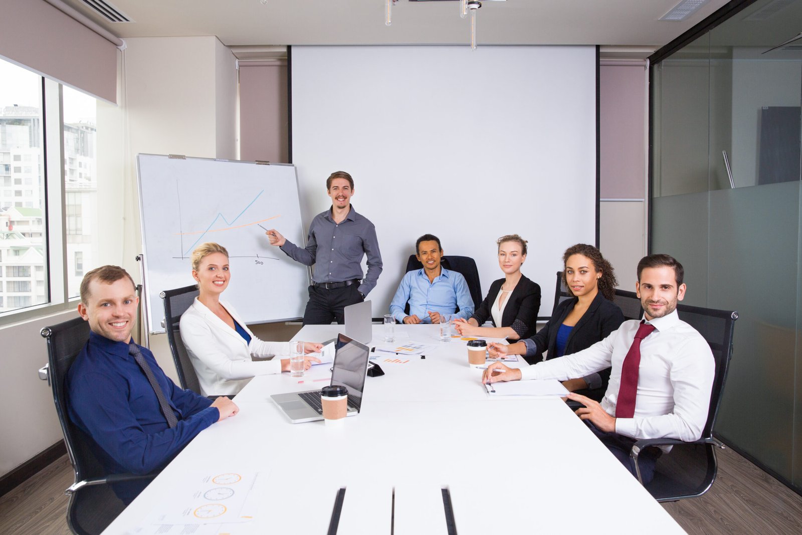 Young businessman standing at whiteboard and pointing to graph with business people sitting at office desk and smiling, looking at camera